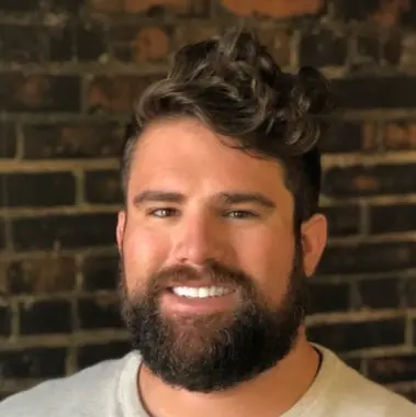 Smiling man with dark curly hair and full beard in front of a brown brick wall.