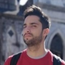 Young man with dark hair and beard wearing a red shirt and backpack outdoors near stone architecture.
