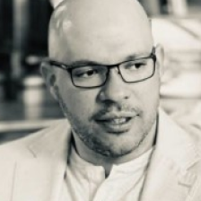 Bald man wearing glasses and a light jacket looking slightly right in a blurred background.