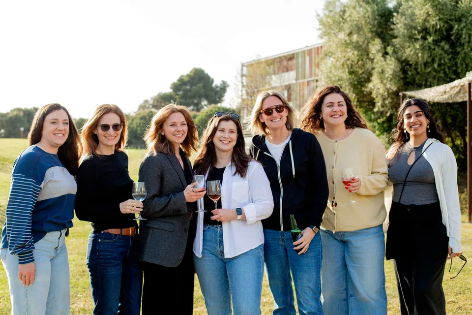 Seven Close teammates smiling side by side on a sunny lawn, several holding glasses of wine